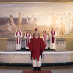 Vihitty punaisessa stoolassa seisoo messuyleisöön päin, takanaan alttarilla piispa avustajineen.