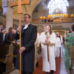 Messukulkue saapuu kirkkoon, ensimmäisenä kulkeva notaari kantaa puista ristiä